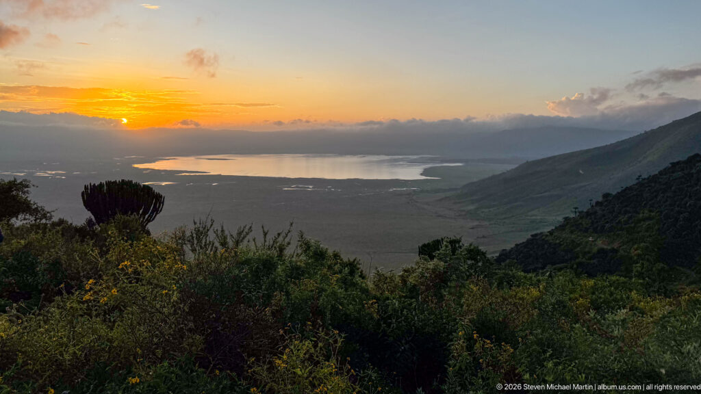 Golden sunset over reflective water on the Ngorongoro Crater floor, framed by dark foreground vegetation and a steep slope.