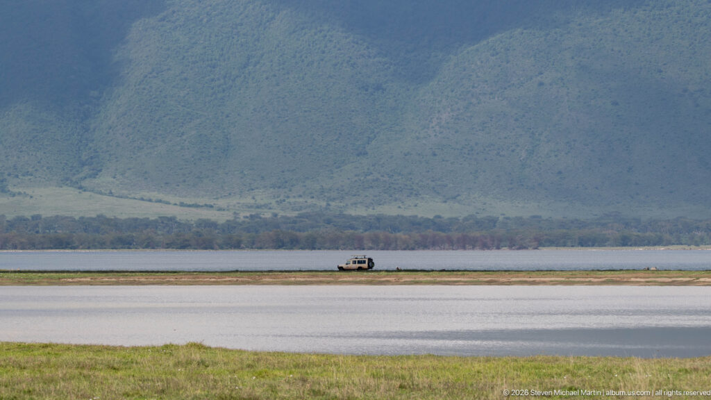 Distant safari vehicle on a narrow track between water bodies beneath the steep green Ngorongoro Crater wall.