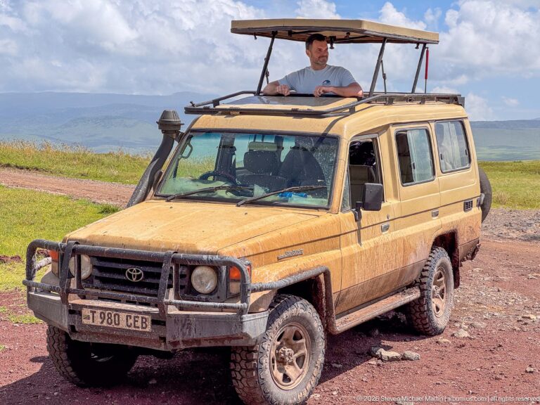 Beige Toyota Land Cruiser with raised safari roof and a man standing through it on the crater floor.