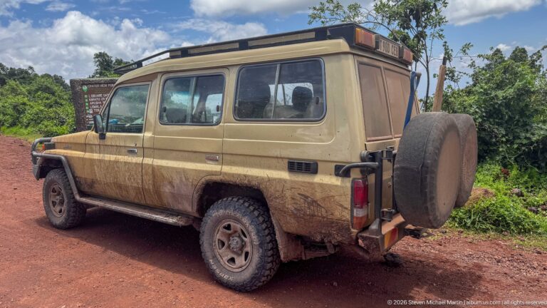 Mud-splashed beige Toyota Land Cruiser safari vehicle parked on a red dirt road beside dense vegetation.