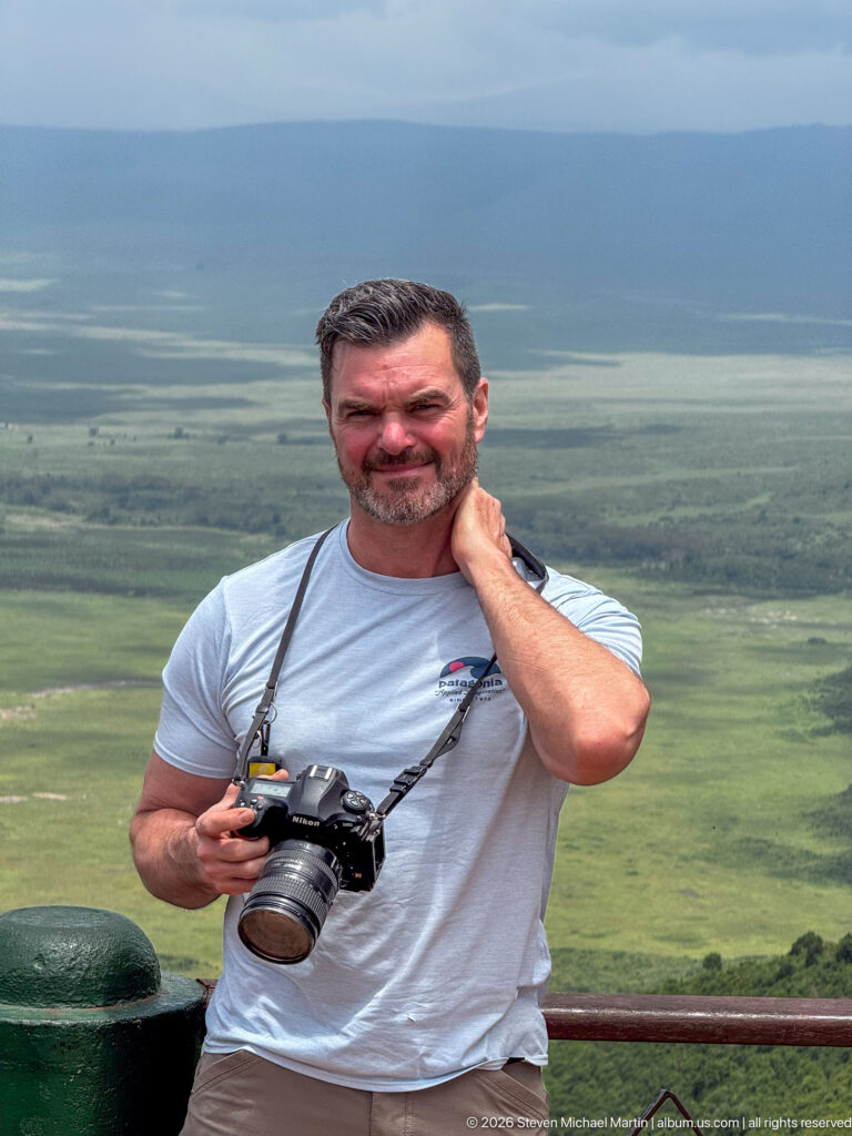 Man holding a camera at a Ngorongoro Crater overlook with the broad green crater floor behind him.