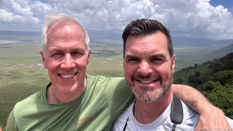 Two smiling men pose at a crater overlook with the green Ngorongoro basin and distant rim behind them.