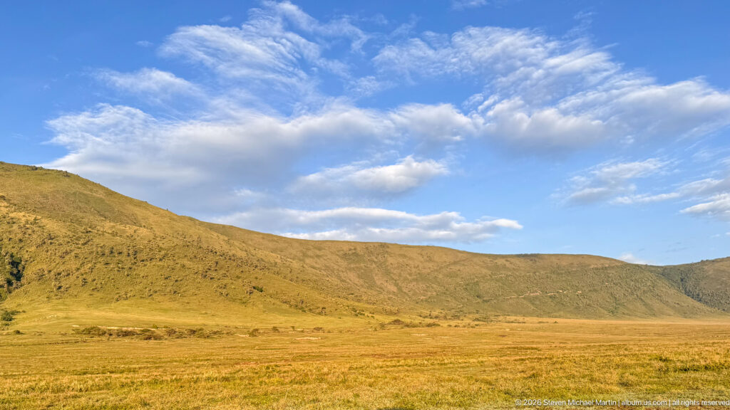 Golden crater grassland beneath the long Ngorongoro wall and a blue sky with scattered white clouds.