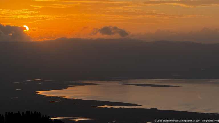 Orange sunset above dark crater ridges and reflective water with irregular shorelines on the Ngorongoro floor.