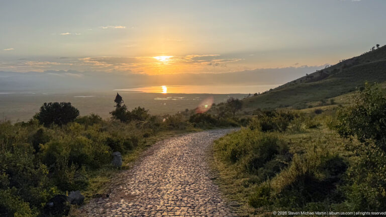 Sunset over Ngorongoro Crater with a cobbled road descending through vegetation toward reflective water on the crater floor.