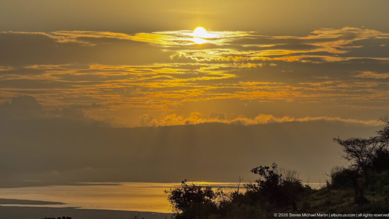 Telephoto sunset over Ngorongoro Crater with golden light reflected on water below silhouetted clouds and vegetation.