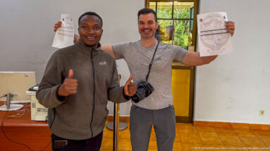 Two smiling men indoors holding Ngorongoro entry documents, one with a camera and both giving thumbs-up gestures.