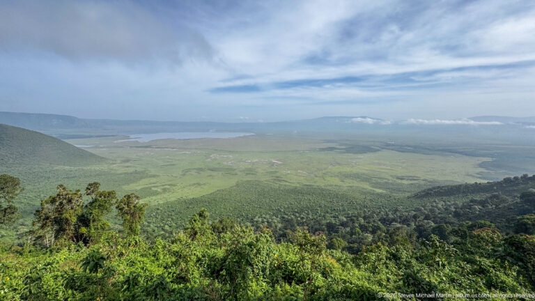 Forested crater rim overlooking a vast green basin and distant lake beneath sweeping white clouds.
