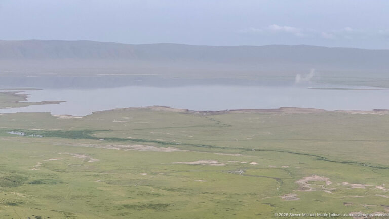 Pale crater-floor lake beyond open grassland, with a hazy escarpment and a small distant vertical plume.