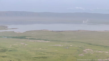 Pale crater-floor lake beyond open grassland, with a hazy escarpment and a small distant vertical plume.
