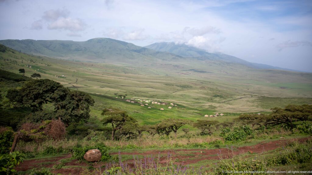 Wide Ngorongoro highland valley with wooded foreground, distant thatched huts, green mountain slopes, and broken cloud.