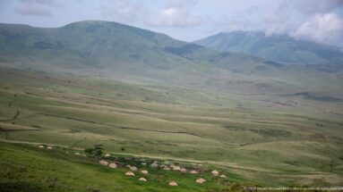 Small cluster of pale thatched huts on broad grassland beneath green mountain ridges and low clouds.
