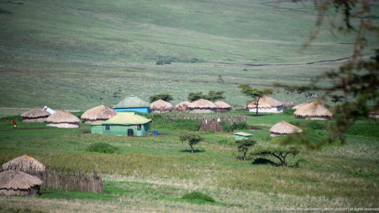 Cluster of round thatched huts around a rectangular pale green-roofed building on a grassy highland slope.