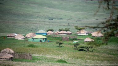 Cluster of round thatched huts around a rectangular pale green-roofed building on a grassy highland slope.