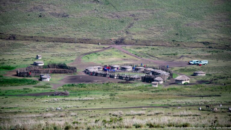 Fenced settlement of round thatched huts beside intersecting dirt tracks, with small figures, livestock, and vehicles.
