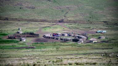 Fenced settlement of round thatched huts beside intersecting dirt tracks, with small figures, livestock, and vehicles.