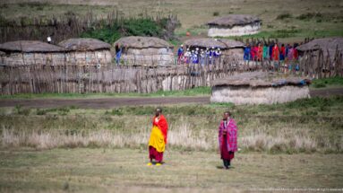 Two foreground figures in bright clothing with a fenced cluster of thatched huts and a group of people behind.