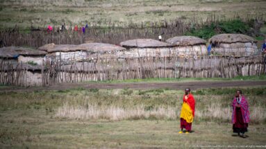 Two people in bright red, yellow, and patterned clothing standing before a stick-fenced settlement with thatched huts.