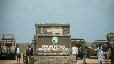 Stone Ngorongoro Conservation Area farewell sign with white rhinoceros silhouette, surrounded by visitors and safari vehicles.