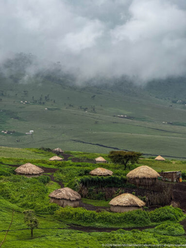 Round thatched huts and fenced pastoral compounds on green Ngorongoro highlands beneath low cloud.