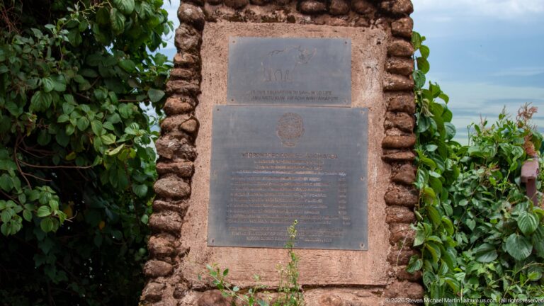 Two metal commemorative plaques mounted vertically in a rough stone monument surrounded by leafy vegetation.