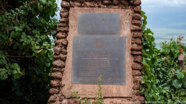 Two metal commemorative plaques mounted vertically in a rough stone monument surrounded by leafy vegetation.