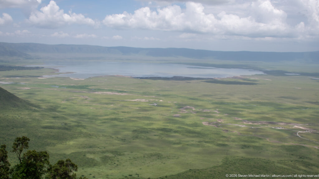 Wide view across green Ngorongoro Crater floor to a broad lake and distant caldera wall under broken clouds.