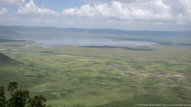Wide view across green Ngorongoro Crater floor to a broad lake and distant caldera wall under broken clouds.