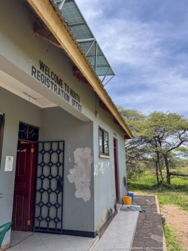 Ndutu Registration Office with painted welcome sign, red doorway, security grille, solar panels, and acacia trees beyond.
