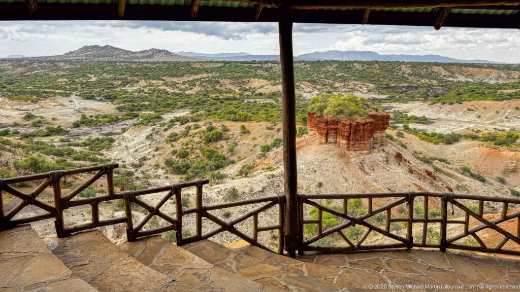 Roofed overlook and timber railing framing a wide view of pale ravines, green scrub, and a red outcrop.