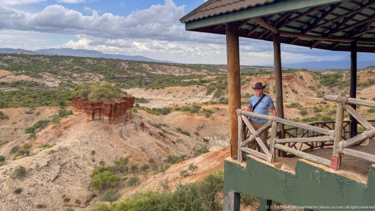 Visitor under a timber-roofed overlook facing a red-brown rock remnant and pale eroded gorge below.
