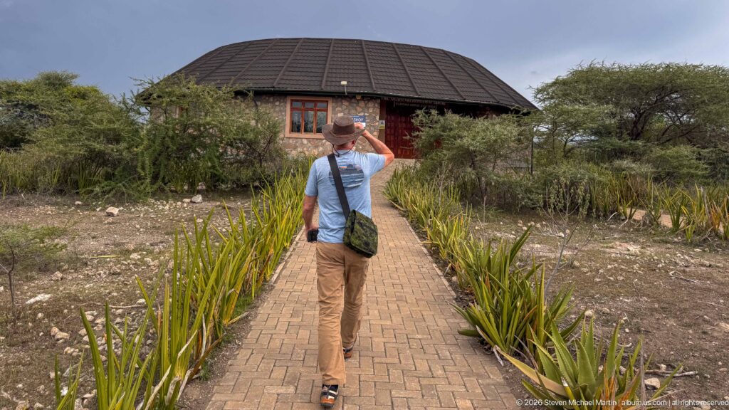 Person walking on a brick path toward a round dark-roofed visitor building surrounded by dry scrub and trees.