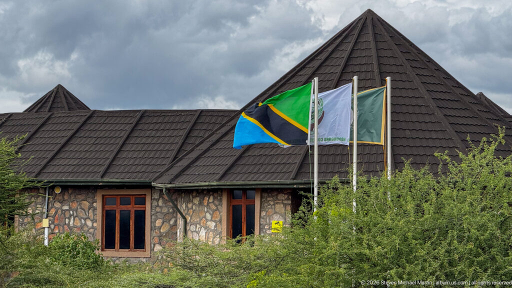 Stone visitor building with steep dark roofs and the Tanzanian flag rising in front amid thorny vegetation.