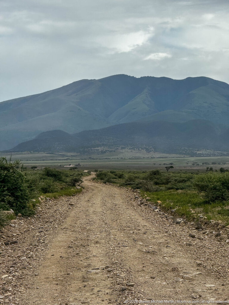 Wide unpaved road crossing green scrub toward dark volcanic ridges beneath an overcast sky.