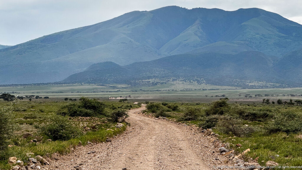 Rocky dirt road through low scrub toward a broad green-gray volcanic mountain under a pale sky.