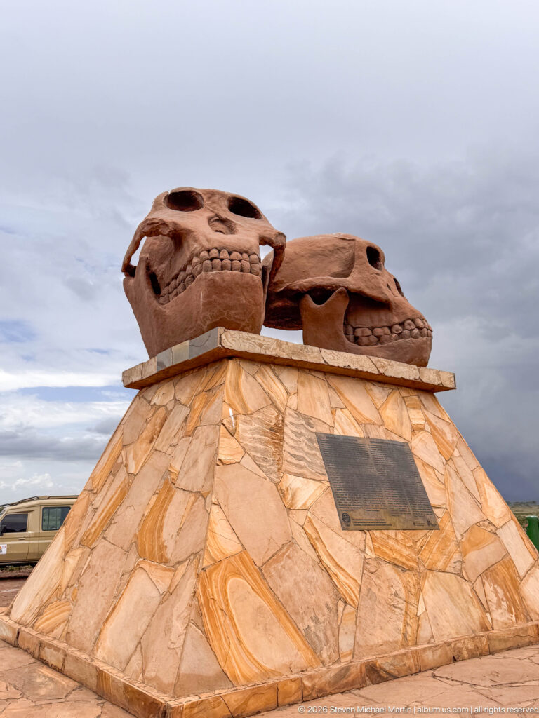 Wide low-angle view of two hominin skull sculptures atop a faceted stone pedestal with plaque.