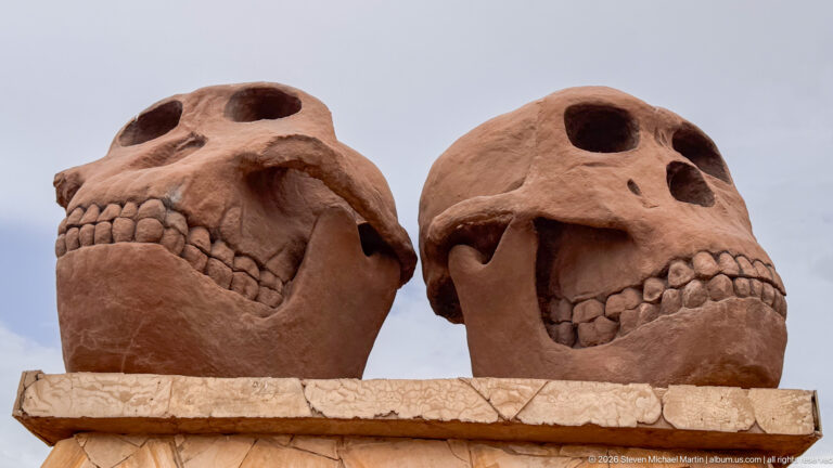 Two monumental reddish hominin skull sculptures mounted side by side on a stone pedestal.
