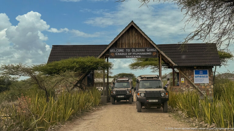 Timber entrance gate reading “Welcome to Olduvai Gorge, Cradle of Humankind, Tanzania” above two safari vehicles.