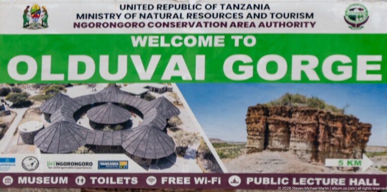 Green-and-white “Welcome to Olduvai Gorge” sign with museum and outcrop images plus visitor-facility icons.