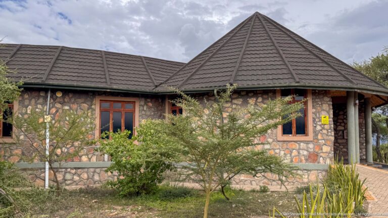 Low stone museum building with wooden windows, cylindrical columns, dark hipped roof, and shrubs in front.