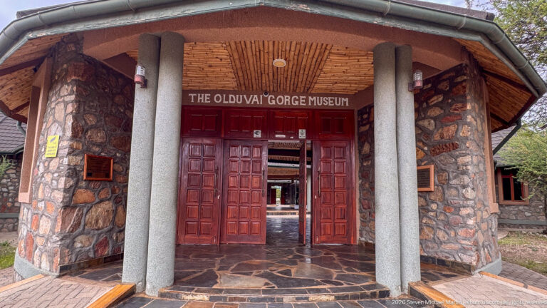 Stone entrance to the Olduvai Gorge Museum with red wooden doors, paired columns, and name above.