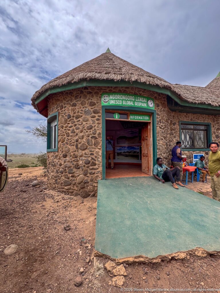 Stone-and-thatch Ngorongoro Lengai UNESCO Global Geopark information building with seated visitors outside.