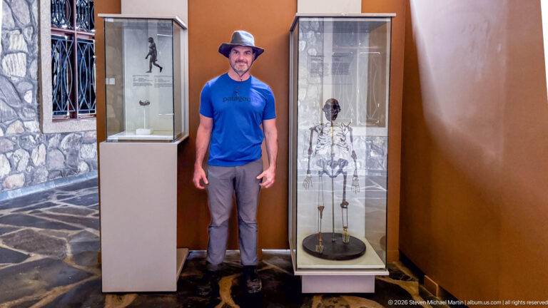 Visitor in blue shirt and hat standing beside a tall glass case containing a mounted hominin skeleton.