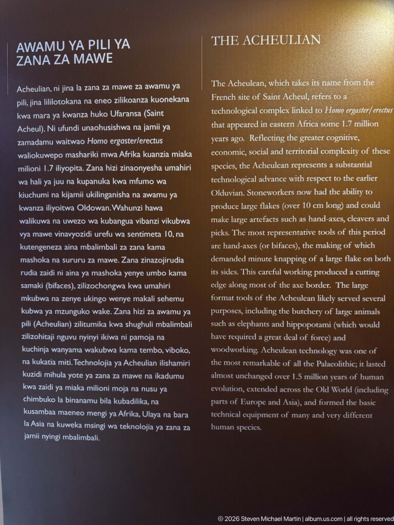 Dark bilingual museum panel titled The Acheulian with explanatory text about the prehistoric stone-tool tradition.