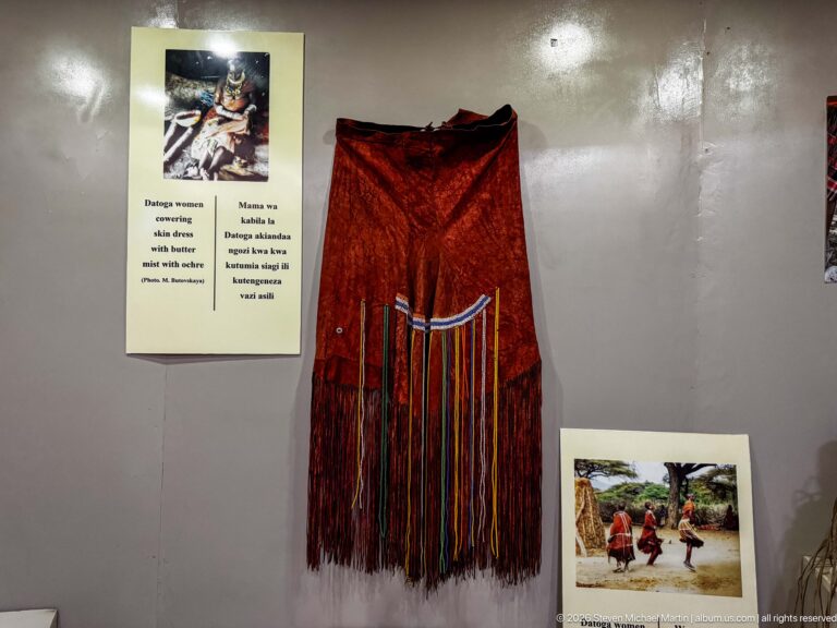 Red hanging Maasai garment with long multicolored fringe and beadwork beside an interpretive label and community photograph.