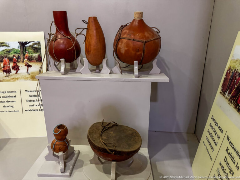 Leather-covered round and elongated containers with straps, plus a small vessel and low lidded container in a museum case.