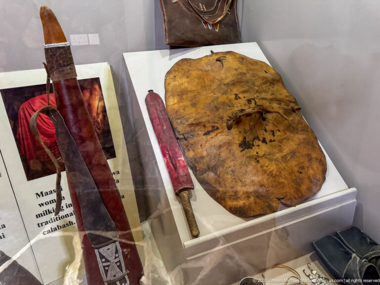 Oval hide shield beside a long dark weapon or sheath in a museum case with Maasai photographs and labels.