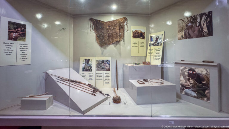 Museum case with long spears or staffs, small ornaments, leatherwork, and photographs relating to Maasai pastoral culture.