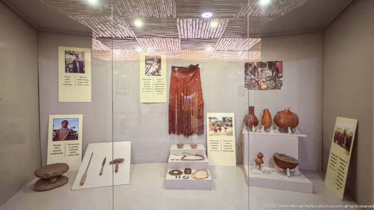 Museum case with a red fringed garment, pottery vessels, small tools, and photographs of Maasai community life.