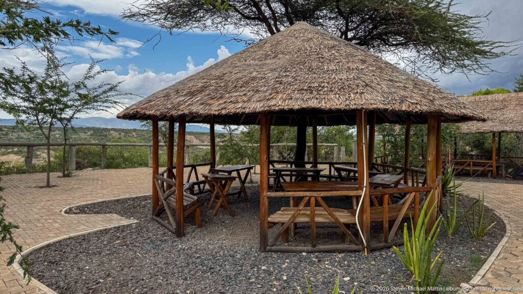 Round timber pavilion with a conical thatched roof, tables, chairs, and open views through surrounding trees.